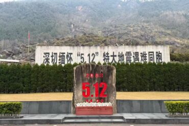 Jiangyou Earthquake Museum,Mianyang,Chinatour Monument with a large red '5.12' and '2008' on a rock in front of a hedge and Chinese text backdrop, mountains in the background (Earthquake memorial).