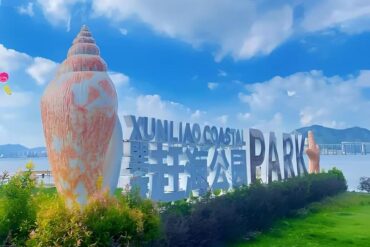 Large white letters spell 'Coastal Park' beside oversized seashell sculptures along a waterfront park, with a green lawn and blue sky in the background.