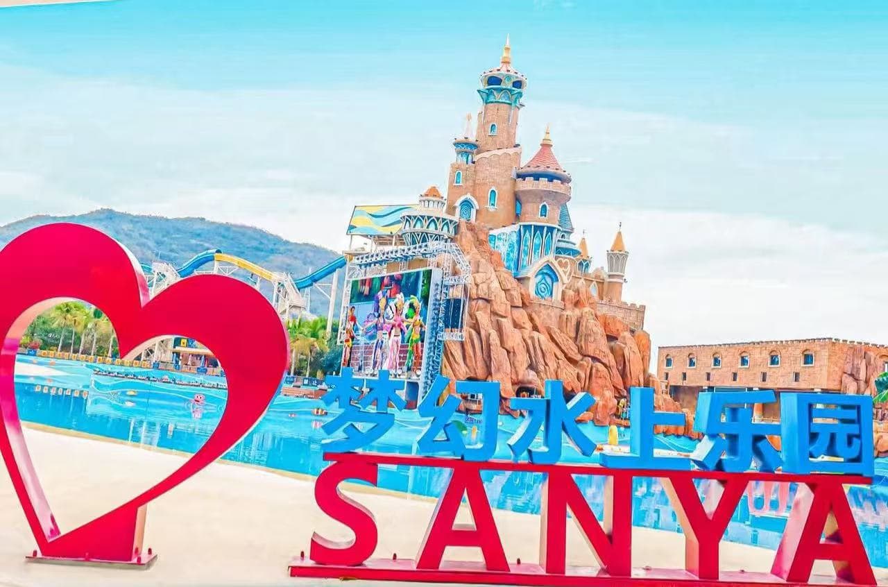 A bright water park entrance with a large pink heart sculpture and blue sign reading 梦幻水上乐园 SANYA against a castle backdrop.
