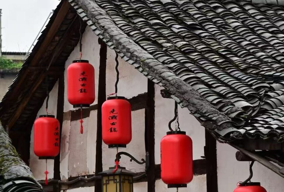 Red cylindrical Chinese lanterns hanging from a traditional tiled roof with white walls and wooden beams, daytime scene