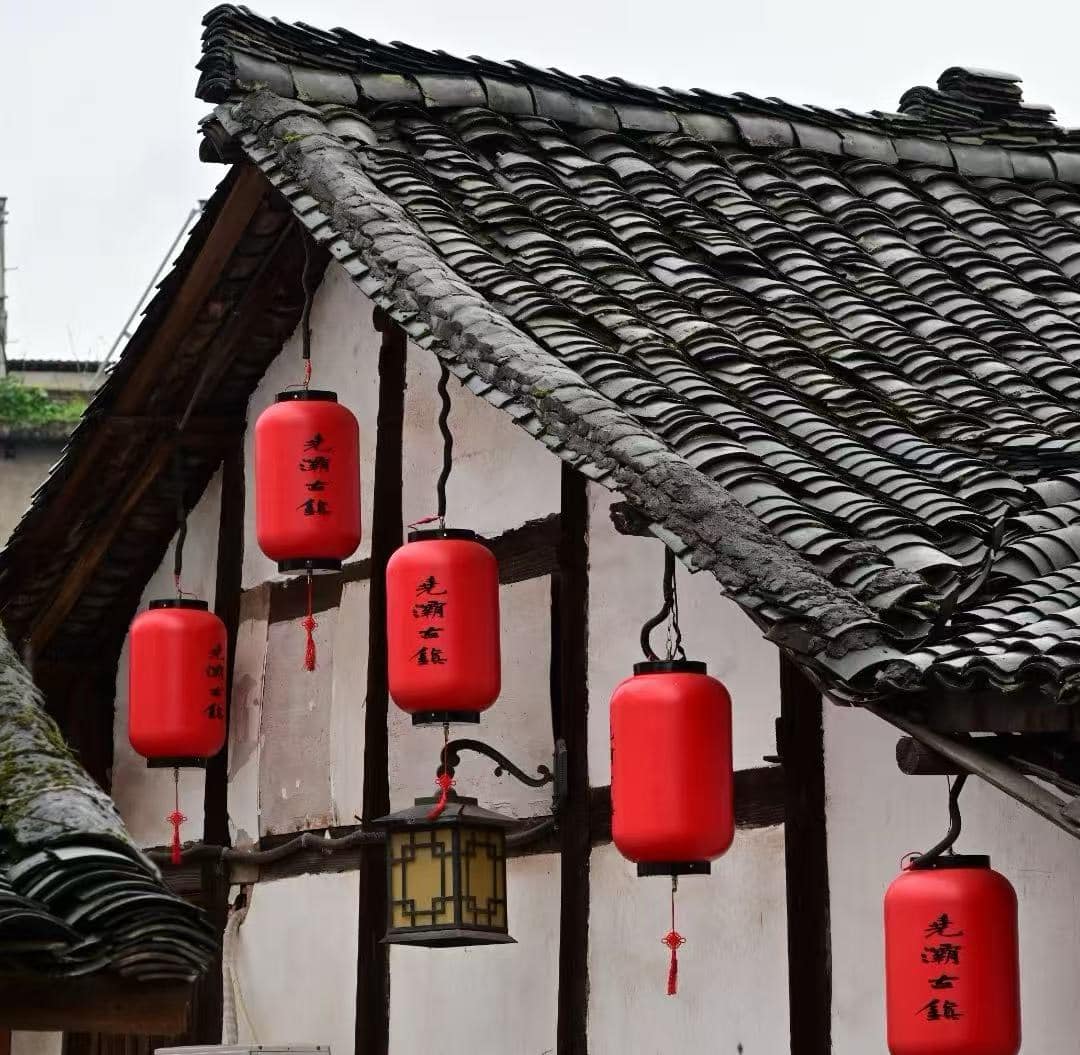 Red cylindrical Chinese lanterns hanging from a traditional tiled roof with white walls and wooden beams, daytime scene