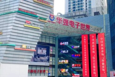 Exterior view of the Huaqiang Electronic World shopping complex with a glass facade, red vertical banners, and colorful brand signs on the building exterior, in a busy urban area.
