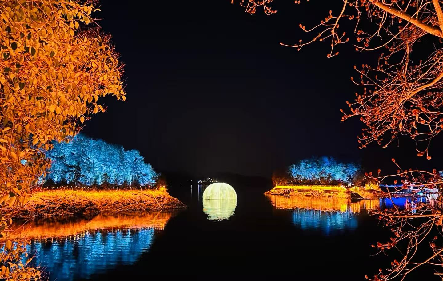Thousand Islands Lake,Hangzhou,Chinatour Night scene of a round illuminated bridge over a calm river, with orange-lit trees along the banks and blue-lit trees in the background reflecting in the water, at night.