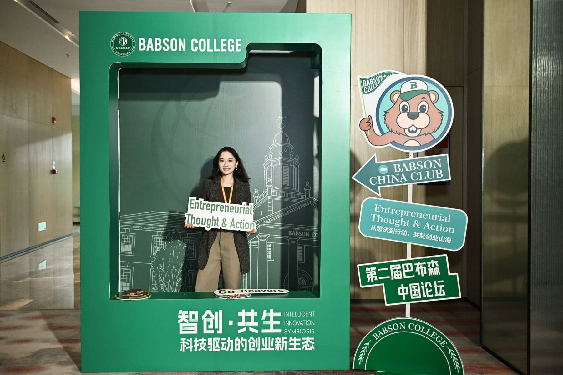 Woman poses inside a large green Babson College photo frame, holding a sign that reads 'Entrepreneurial Thought & Action' amid campus graphics and mascot signs.