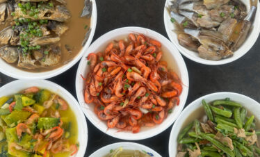 Assortment of cooked seafood dishes in white bowls: vibrant shrimp in the center with fish pieces and mixed vegetables around it.