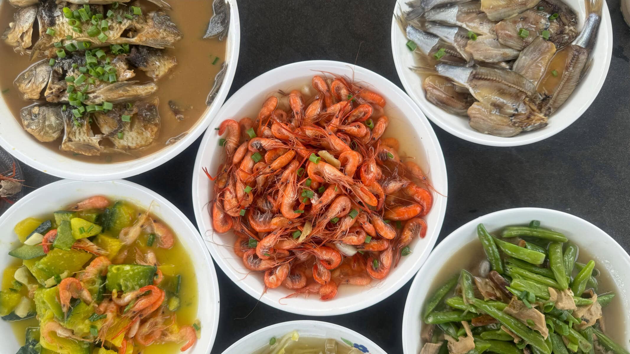 Assortment of cooked seafood dishes in white bowls: vibrant shrimp in the center with fish pieces and mixed vegetables around it.