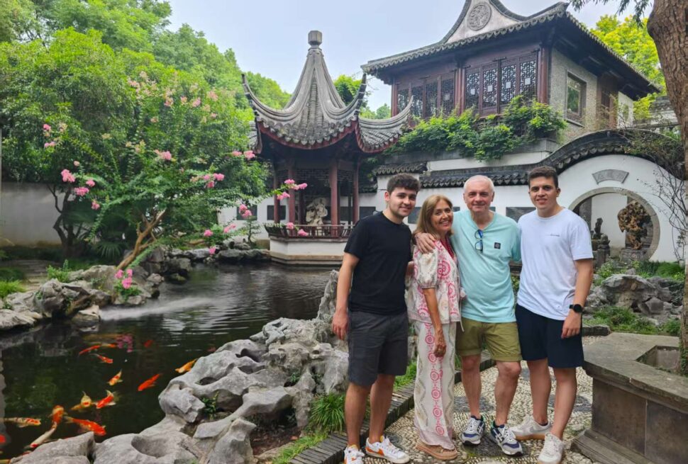 Four people pose together in a traditional Chinese garden with koi pond and a curved pavilion behind them.