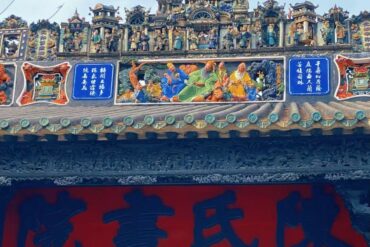 Ornate temple roof with colorful carvings and figural sculptures, blue plaques with Chinese characters, and a large red banner beneath.