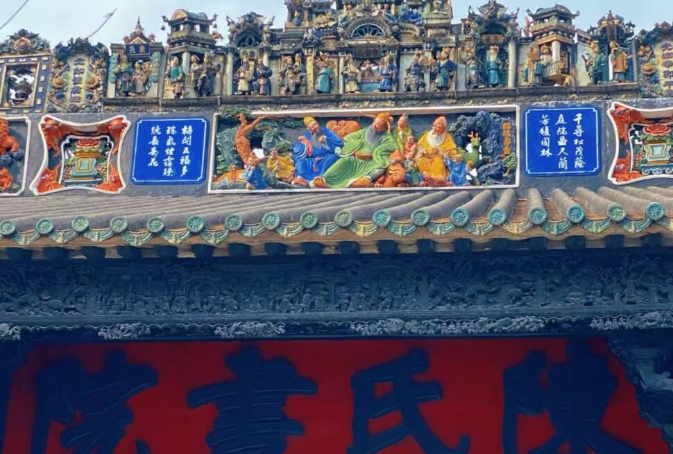 Chen Clan Ancestral Hall,Guangzhou,Chinatour Ornate temple roof with colorful carvings and figural sculptures, blue plaques with Chinese characters, and a large red banner beneath.