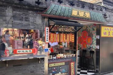 A small Chinese street-side shop selling snacks with hanging lanterns and storefront signs; people line up at the counter.