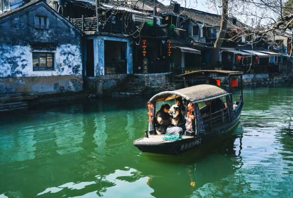 Xitang Water Town,Hangzhou,Chinatour Tour boat with a woven canopy glides along a green canal beside blue-washed houses adorned with red lanterns. Family passengers sit inside the boat.