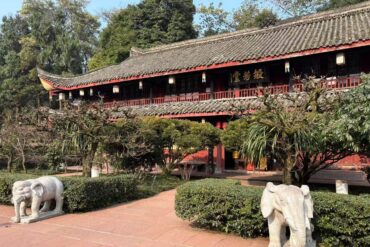 Wannian Temple,Leshan,Chinatour Traditional Chinese temple or pavilion with a tiled roof, red walls, and a signboard, set in a landscaped garden with trimmed hedges and trees; stone elephant statues on the path in the foreground