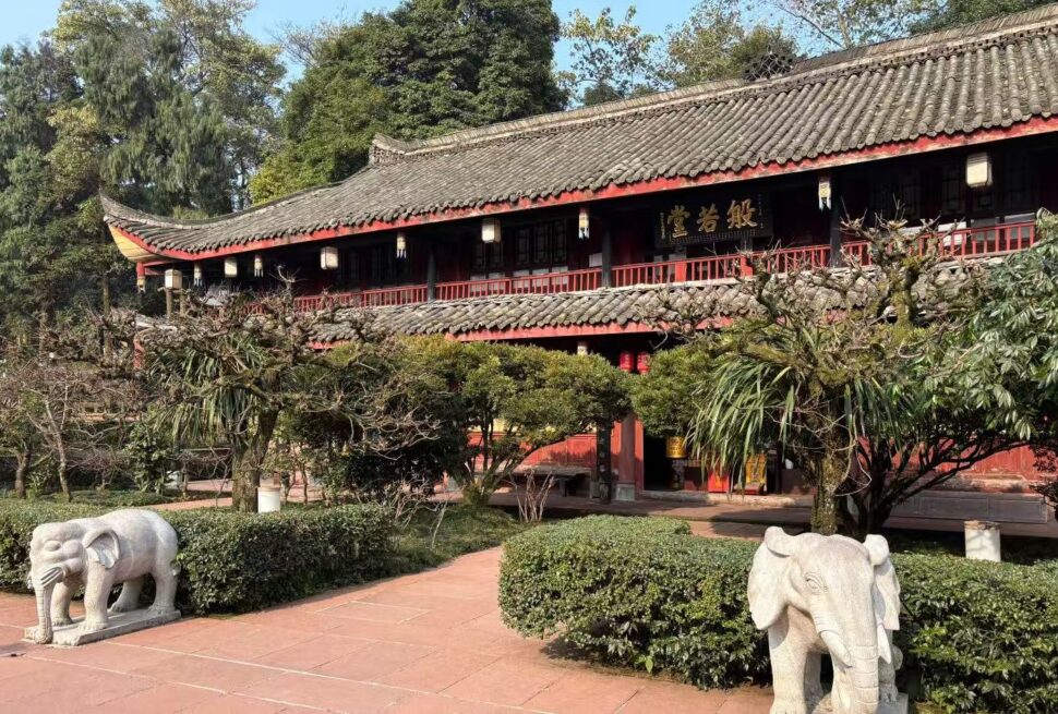 Wannian Temple,Leshan,Chinatour Traditional Chinese temple or pavilion with a tiled roof, red walls, and a signboard, set in a landscaped garden with trimmed hedges and trees; stone elephant statues on the path in the foreground