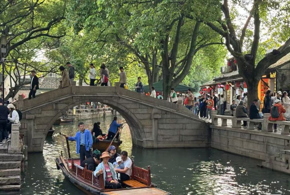 Tongli Water Town,Suzhou,Chinatour Stone arched pedestrian bridge over a canal with people walking; boats on the water and trees overhead on a sunny day.