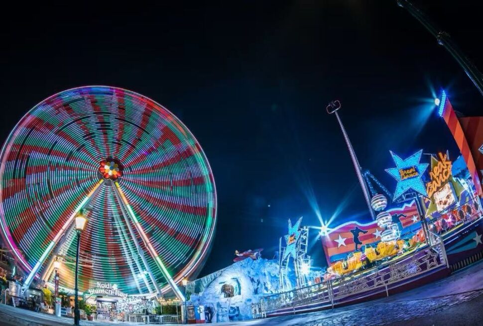 Happy Valley,Shenzhen,Chinatour Nighttime carnival scene with a brightly lit spinning Ferris wheel and colorful rides along a illuminated promenade at a fairground edge nearby edges of the park.