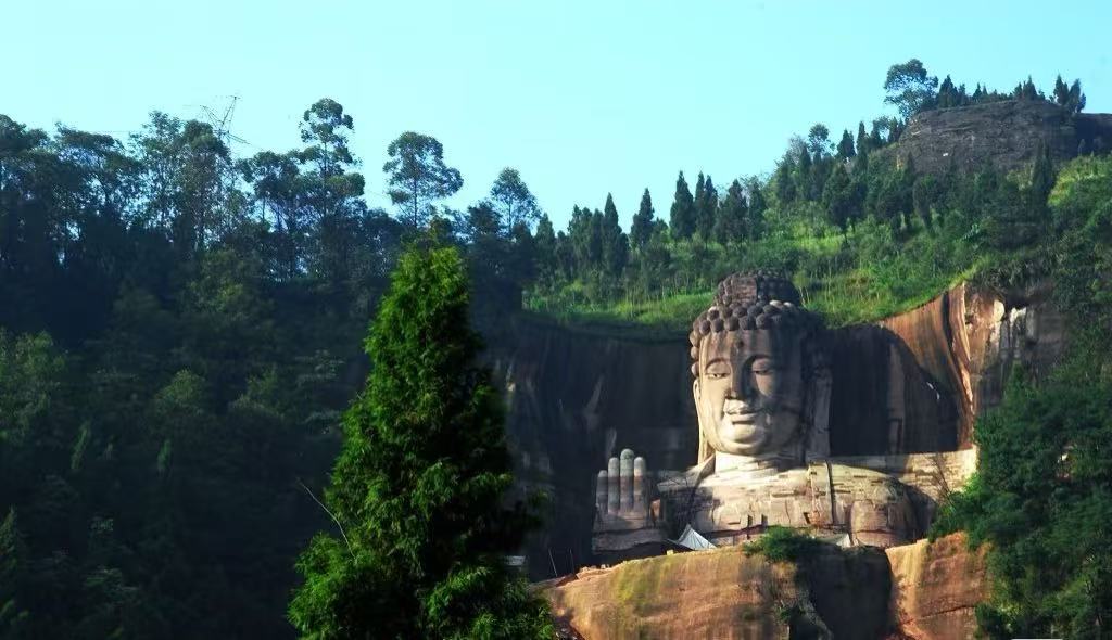 Lingyun Mountain National Forest Park,Leshan,Chinatour Large carved Buddha statue in a cliff face, with right hand raised in blessing, surrounded by trees.