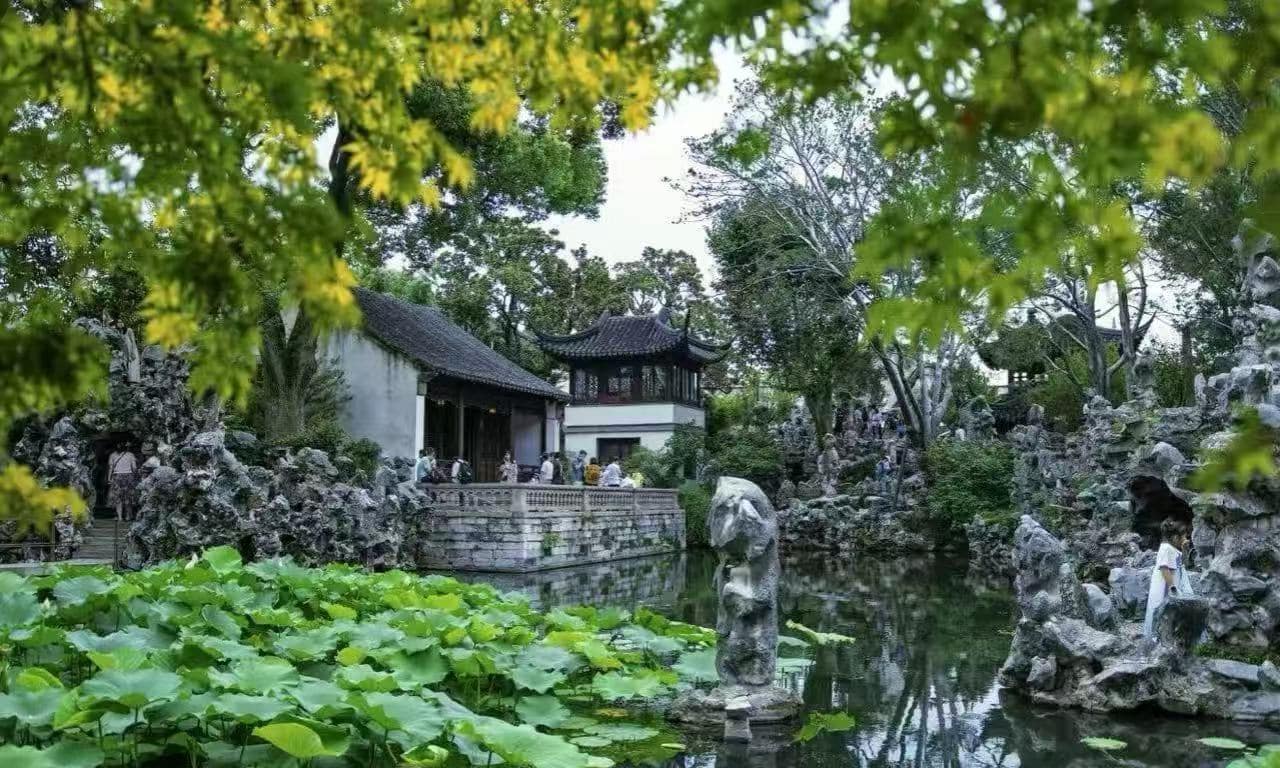 Lion Grove Garden,Suzhou,Chinatour People walk along a stone terrace beside a pond in a traditional Chinese garden with rock formations and a pagoda-style pavilion.