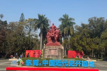 Guangdong Opium War Museum,Dongguan,Chinatour Red statues group with a cannon on a stone pedestal in a park, palm trees behind, blue sky above, bold blue Chinese lettering in front.