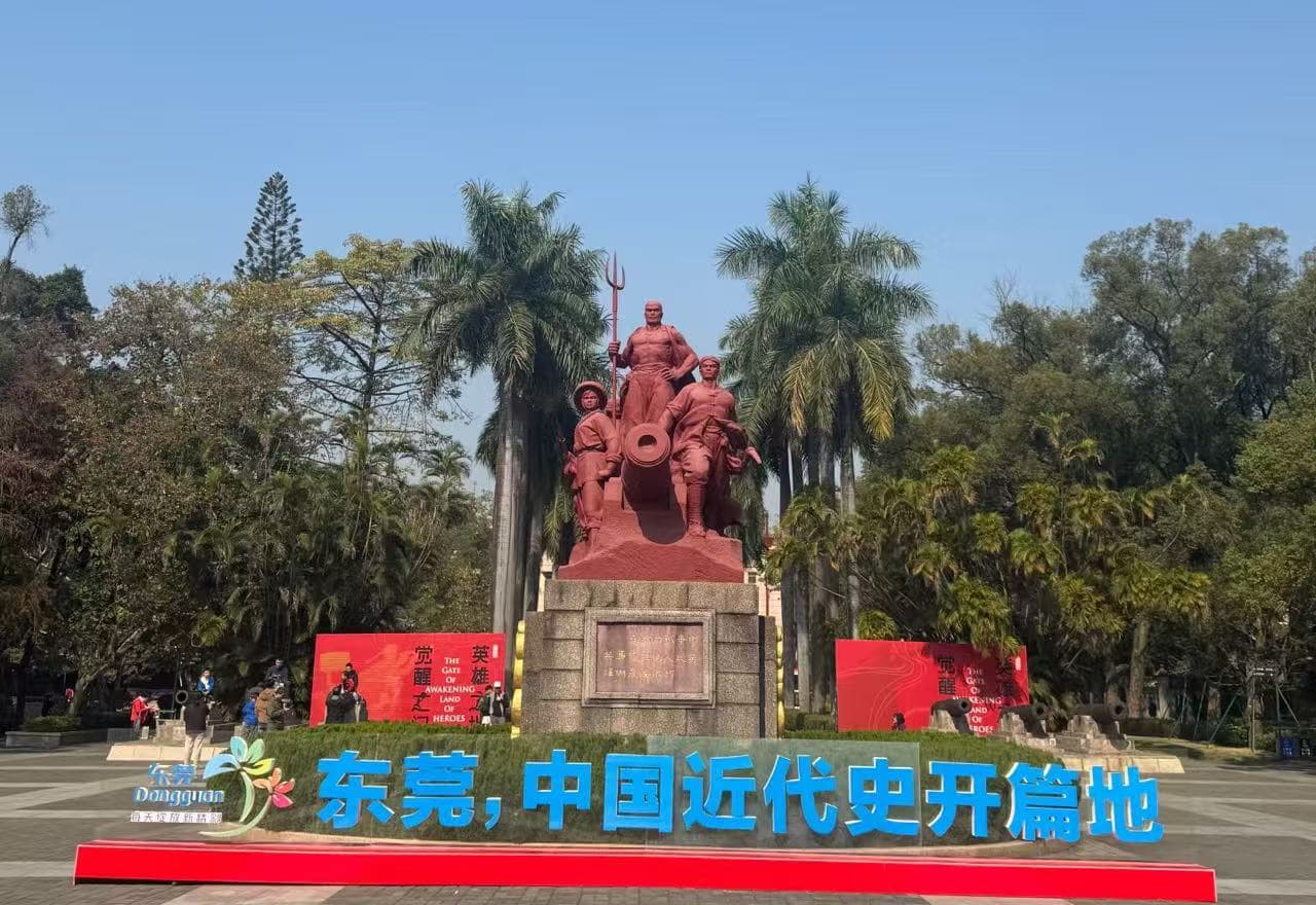 Guangdong Opium War Museum,Dongguan,Chinatour Red statues group with a cannon on a stone pedestal in a park, palm trees behind, blue sky above, bold blue Chinese lettering in front.