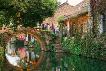 Zhouzhuang Water Town,Suzhou,Chinatour Stone arch bridge over a green canal, ivy-covered walls, and people strolling along a historic riverside walkway.