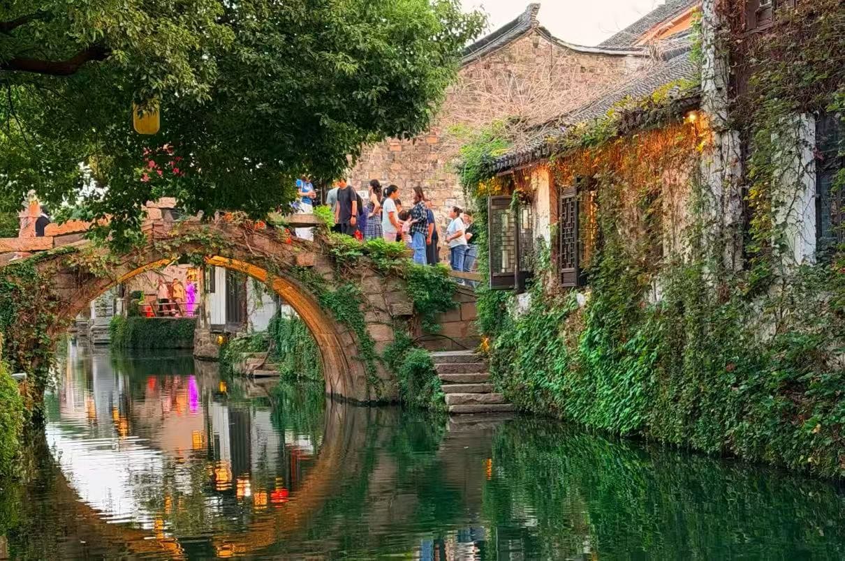 Zhouzhuang Water Town,Suzhou,Chinatour Stone arch bridge over a green canal, ivy-covered walls, and people strolling along a historic riverside walkway.