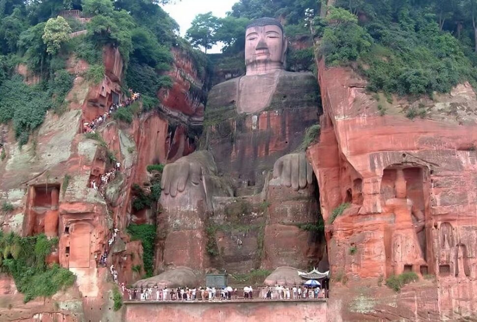 Leshan Giant Buddha,Leshan,Chinatour Giant Buddha statue carved into a red cliff with a calm face and large hands, surrounded by greenery and visitors along a stair path on the left.