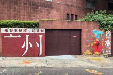 Red wall with large white Chinese characters beside a brown double gate; right side features a colorful mural with a water faucet and gold ingots.