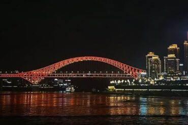Chaotianmen Dock and Riverside Promenade,Chongqing,Chinatour Nighttime view of a red-lit arched bridge spanning a river with a city skyline to the right; reflections glow on the water.