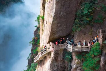 Xihai Grand Canyon,Huangshan,Chinatour Visitors wearing orange helmets walk along a railinged cliff path carved into the rocky mountainside beside a misty waterfall to the left.