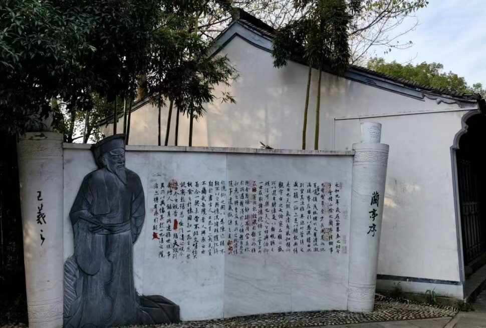 Carved stone relief of a seated scholar beside a wall with Chinese calligraphy and columns in a garden setting.