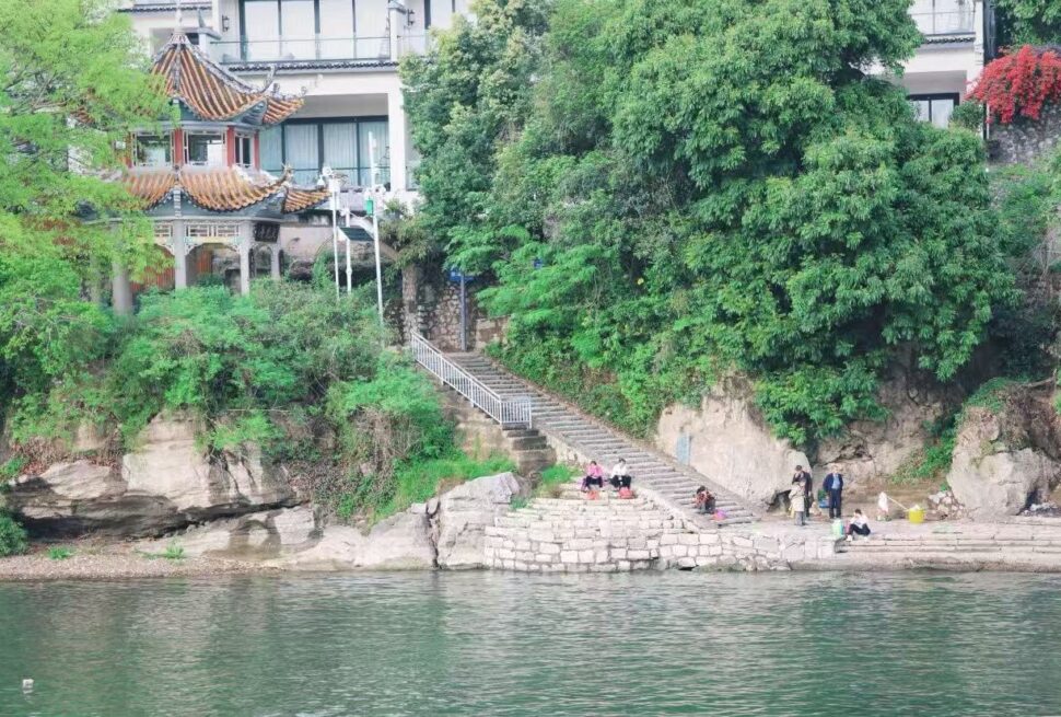 People sit and relax on stone steps along a riverbank, with green trees and a traditional tiled pavilion in the background.Fuli Ancient Town,Yangshuo,Guangxi,Chinatravel