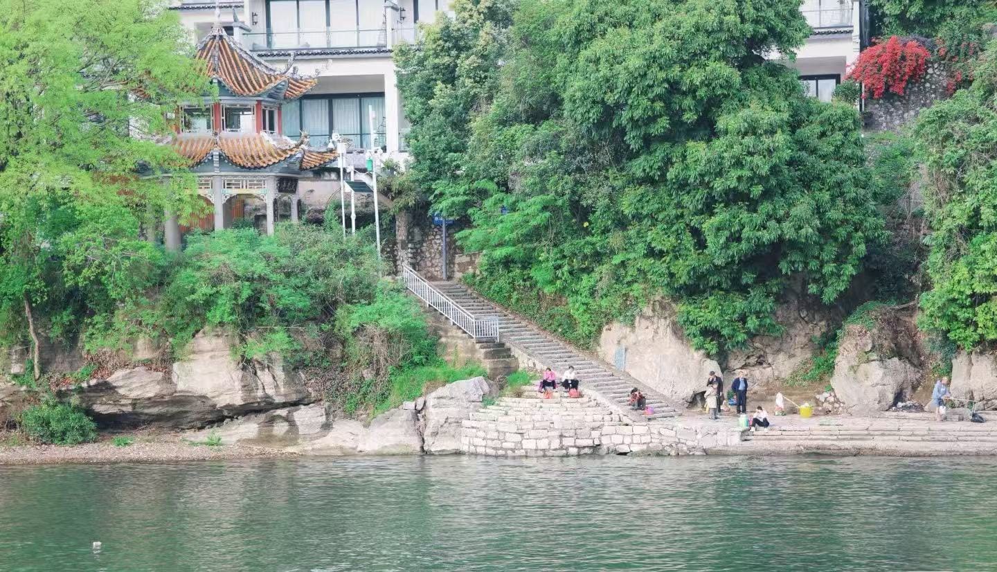 People sit and relax on stone steps along a riverbank, with green trees and a traditional tiled pavilion in the background.Fuli Ancient Town,Yangshuo,Guangxi,Chinatravel