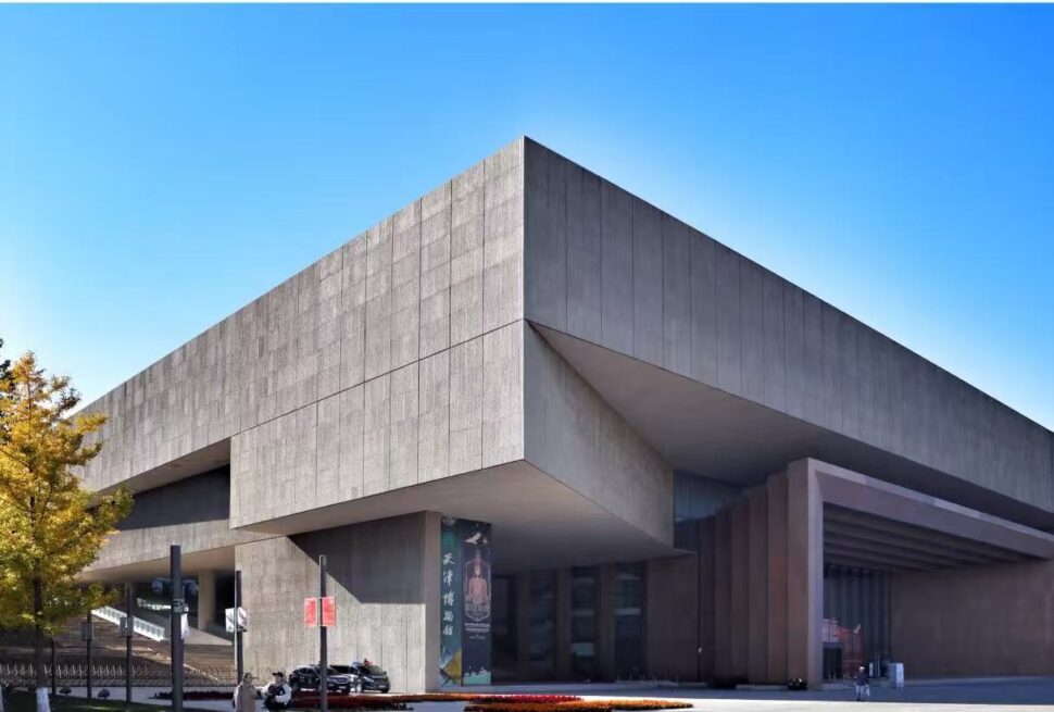 Tianjin Museum,Tianjin,Chinatour Large modern concrete building with a cantilevered upper section, glass entrance, and autumn trees on the left under a clear blue sky.