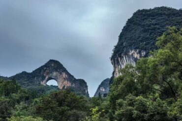 Scenic forested valley with a natural stone arch in the left hill under a cloudy sky.Moon Hill,Yangshuo,Guangxi,Chinatravel
