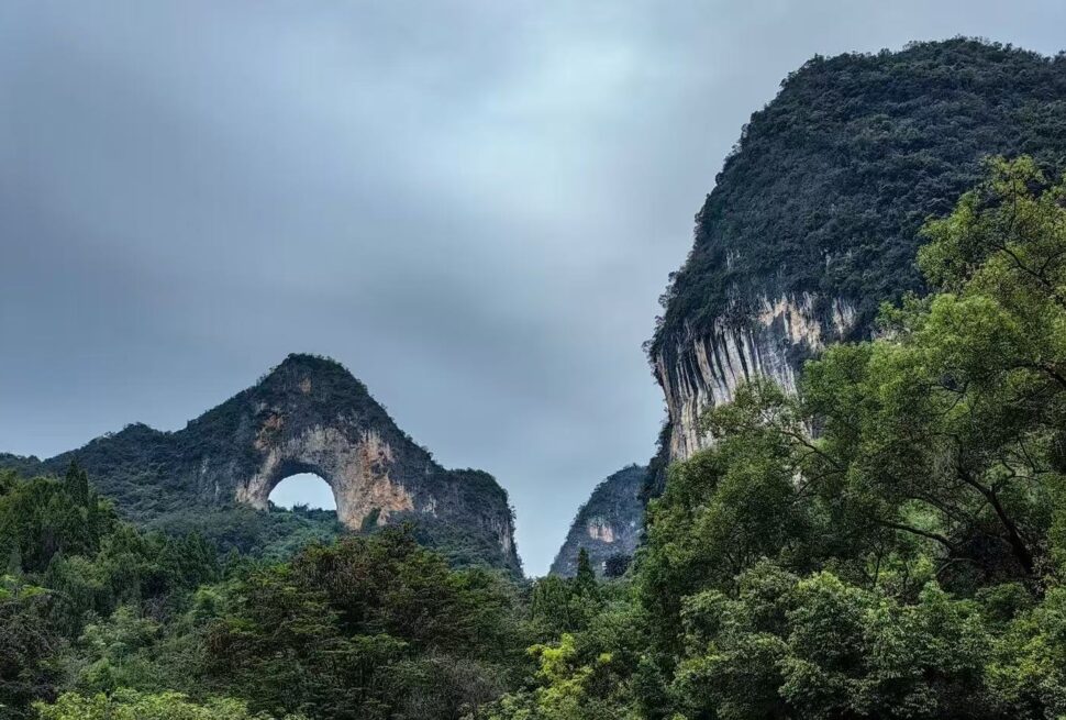 Scenic forested valley with a natural stone arch in the left hill under a cloudy sky.Moon Hill,Yangshuo,Guangxi,Chinatravel