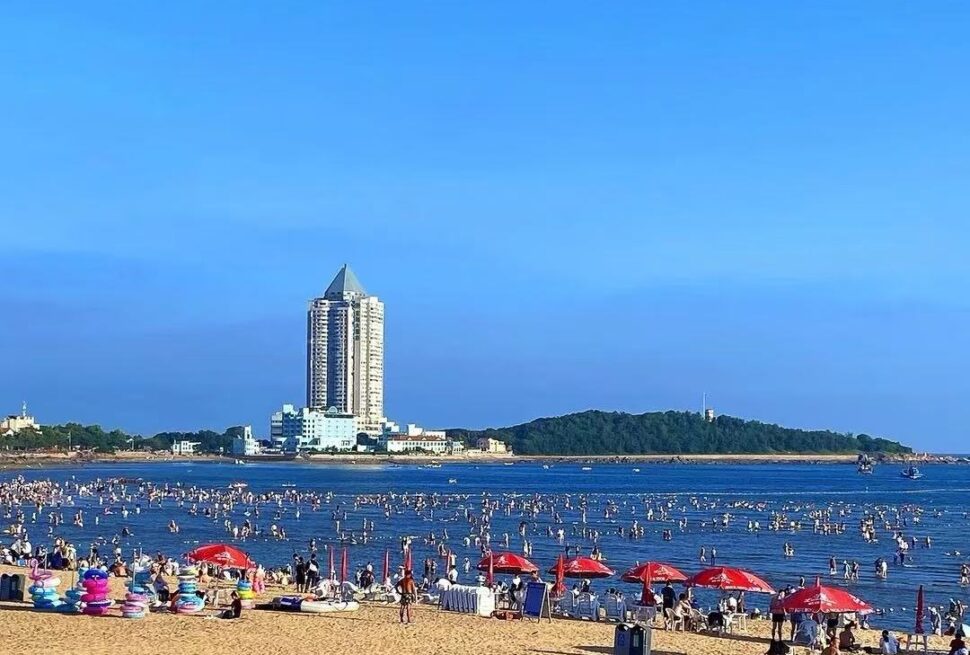 Crowded beach with people in shallow water, red umbrellas on the sand, and a tall pyramid-topped building in the distance against a blue sky.