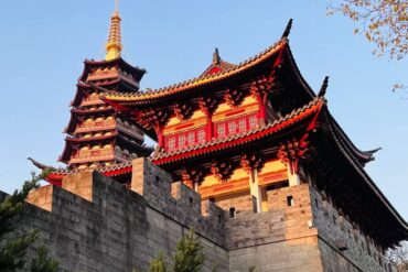 Wuzhou Ancient City,Jinhua,Chinatour Red and black multi-tiered Chinese pagoda atop a stone fortress wall against a clear blue sky, with ornate curved roofs and golden pinnacle.
