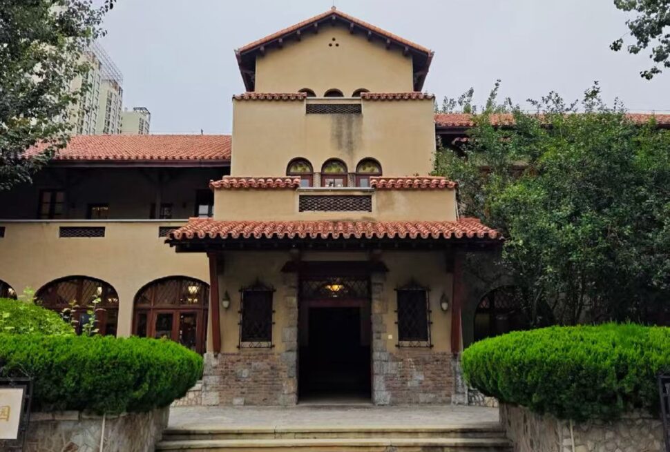Villa-style building with red-tiled roof, arched windows, and a central entrance framed by trimmed hedges in a garden setting.