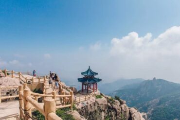 Mount Laoshan Scenic Area ,Qingdao,Chinatour Hikers walk along a wooden railing on a rocky mountain path, with a small blue-roofed pavilion perched on the rocks.