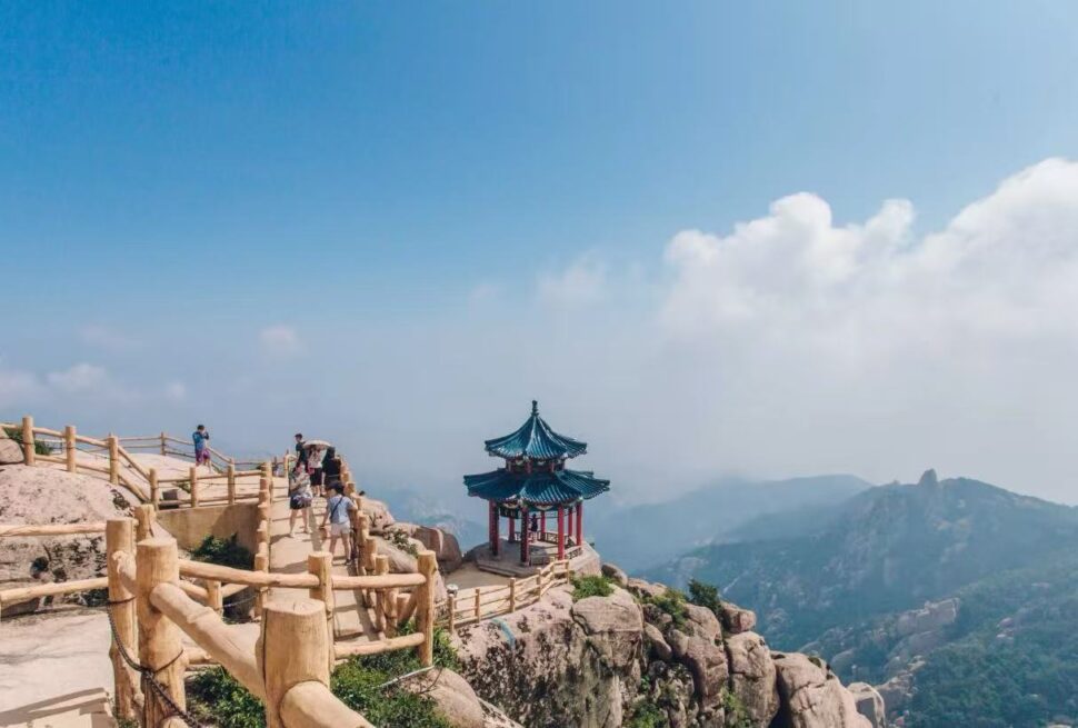 Mount Laoshan Scenic Area ,Qingdao,Chinatour Hikers walk along a wooden railing on a rocky mountain path, with a small blue-roofed pavilion perched on the rocks.