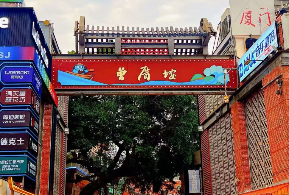 Zengcuoan Coastal Art Village,Xiamen,Chinatour Decorative red gate over a street with Chinese characters, flanked by colorful signs; cartoon penguin in a blue boat on the left.