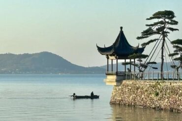 Dongqian Lake,Ningbo,Chinatour Lakeside scene with a pagoda-style pavilion on a stone platform and two people rowing a small boat on calm water, hills in the distance.