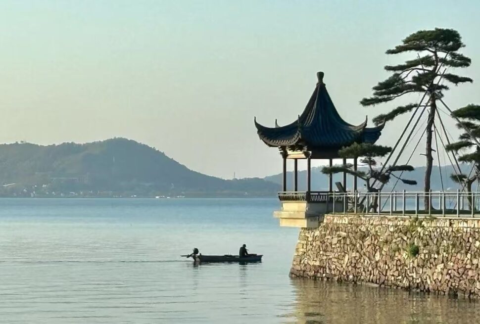 Dongqian Lake,Ningbo,Chinatour Lakeside scene with a pagoda-style pavilion on a stone platform and two people rowing a small boat on calm water, hills in the distance.