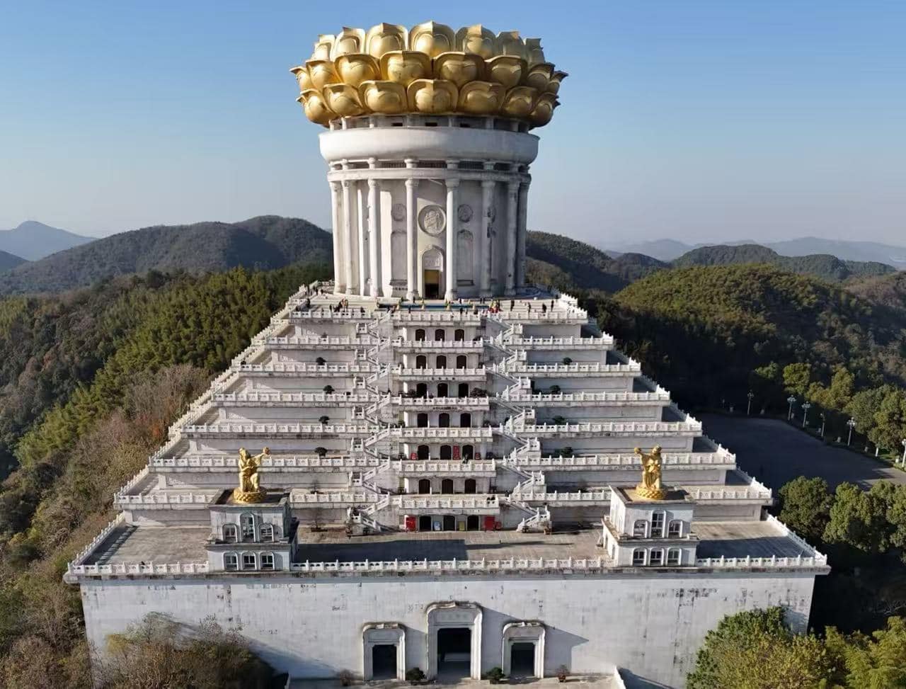A giant white tiered temple on a hill, topped with a gilded lotus crown and surrounded by forested mountains.