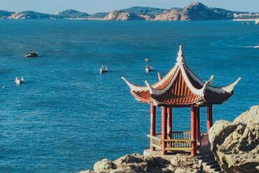 Red-roofed Chinese pavilion on rocky coast overlooking a calm blue sea with distant boats and islands in the background,Meizhou Island,Putian,Fujian,Chinatravel
