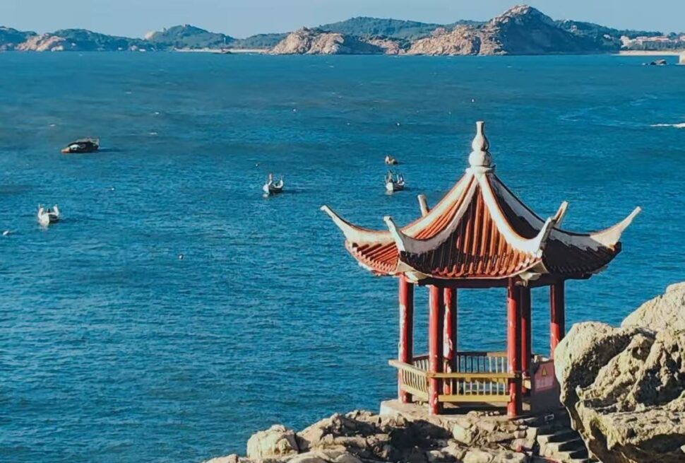 Red-roofed Chinese pavilion on rocky coast overlooking a calm blue sea with distant boats and islands in the background,Meizhou Island,Putian,Fujian,Chinatravel