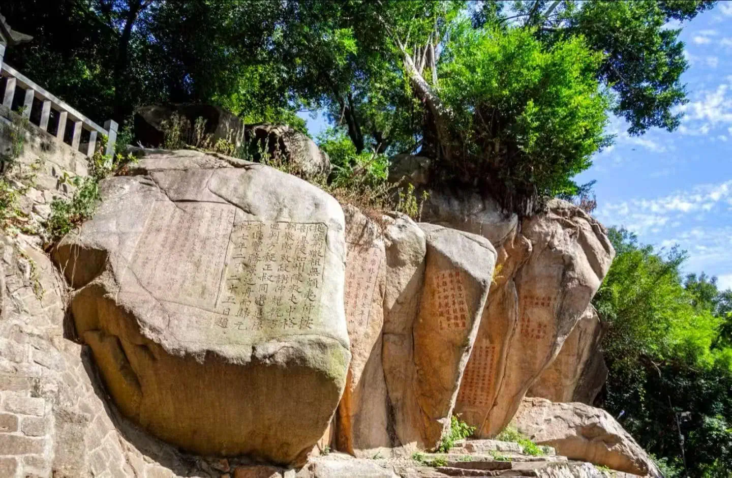 Large boulder with Chinese inscriptions carved on its face, set among rocks and trees on a sunny hillside.