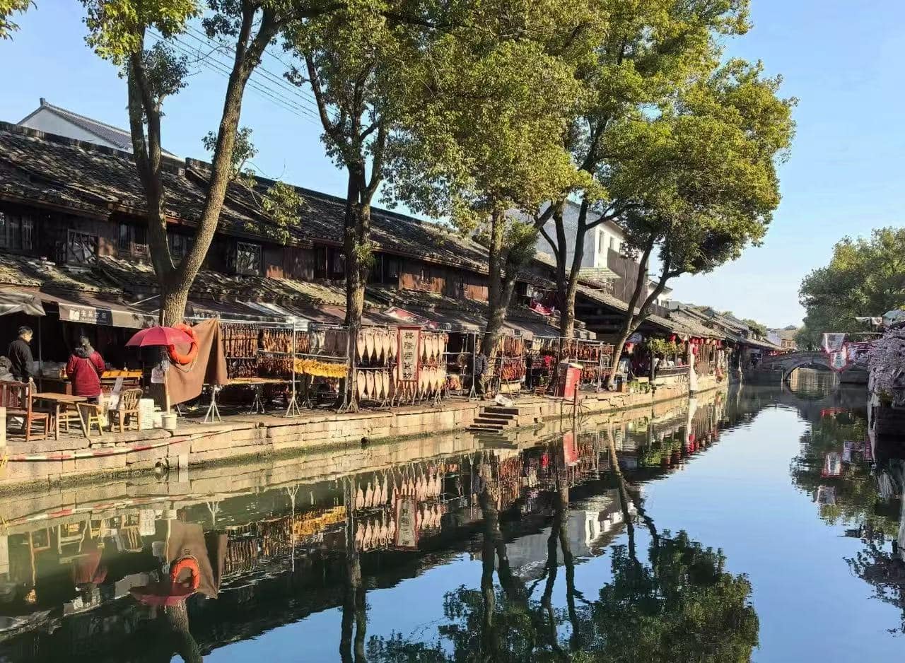 Canal-side street with traditional buildings, market stalls, and trees, reflected in calm water beneath a clear blue sky.