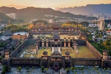 Hengdian World Studios ,Jinhua,Chinatour Sprawling ancient Chinese fortress with tall stone walls, pagoda roofs, and inner courtyards at sunset; city and hills in the background.