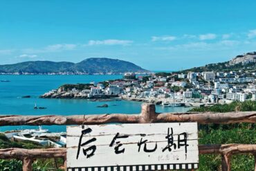 Dongji Island,Zhoushan,Chinatour Coastal town perched on hills with blue harbor water, seen behind a rustic wooden railing and a white sign with Chinese characters in the foreground.